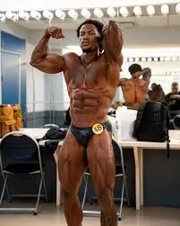 Christian Swole Ricketts posing backstage in a bodybuilding/fitness setting, flexing his muscles in front of a mirror an